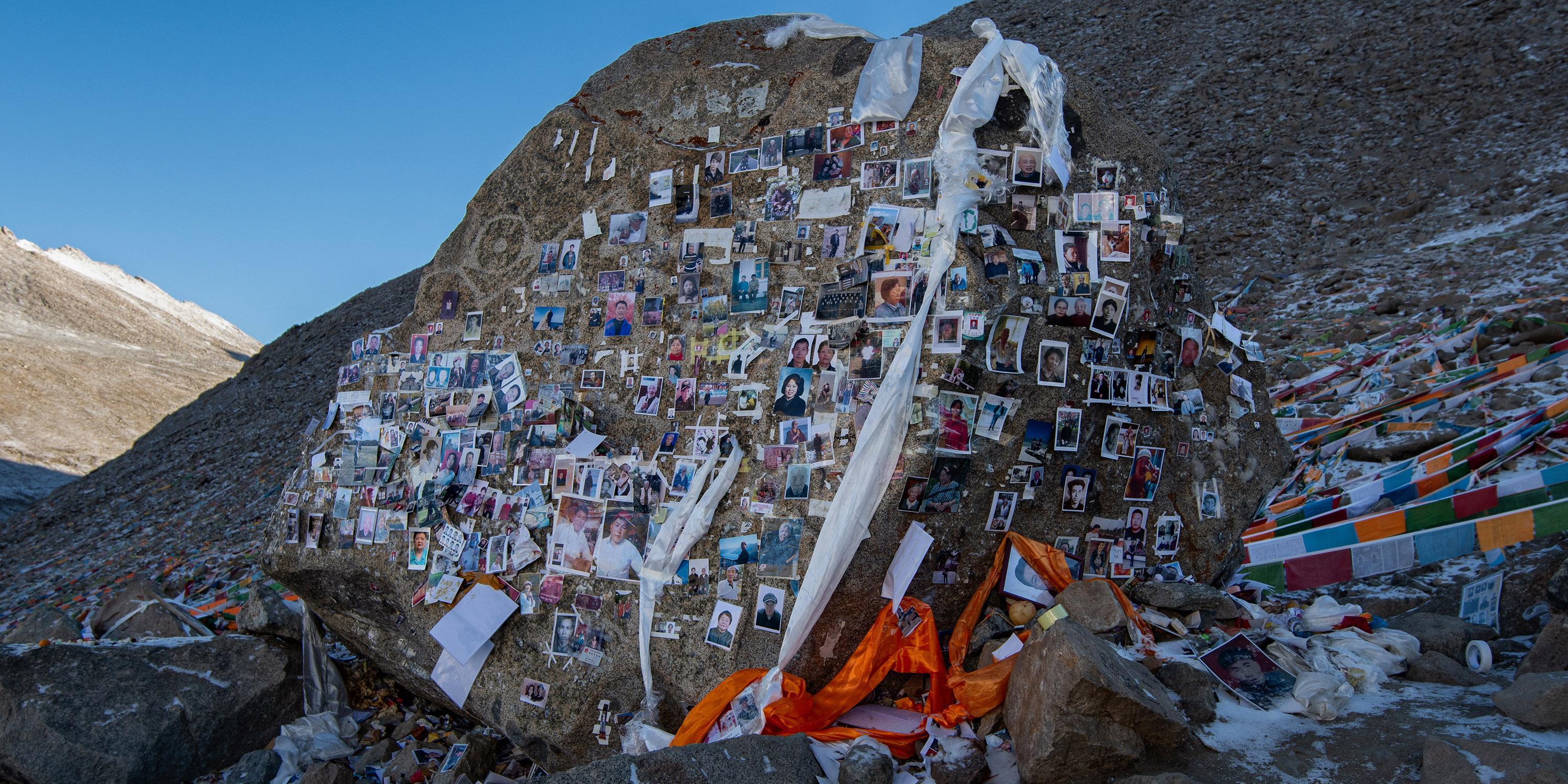 A large rock on a mountain slope covered with memorial photographs and draped with prayer scarves.