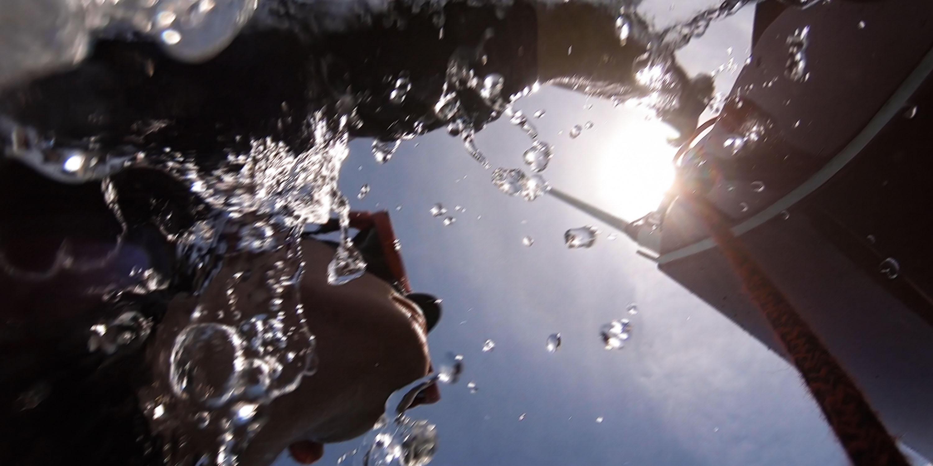 Underwater view of a person swimming near the surface, with sunlight and water droplets caught in motion, while part of a sailboat is visible above.