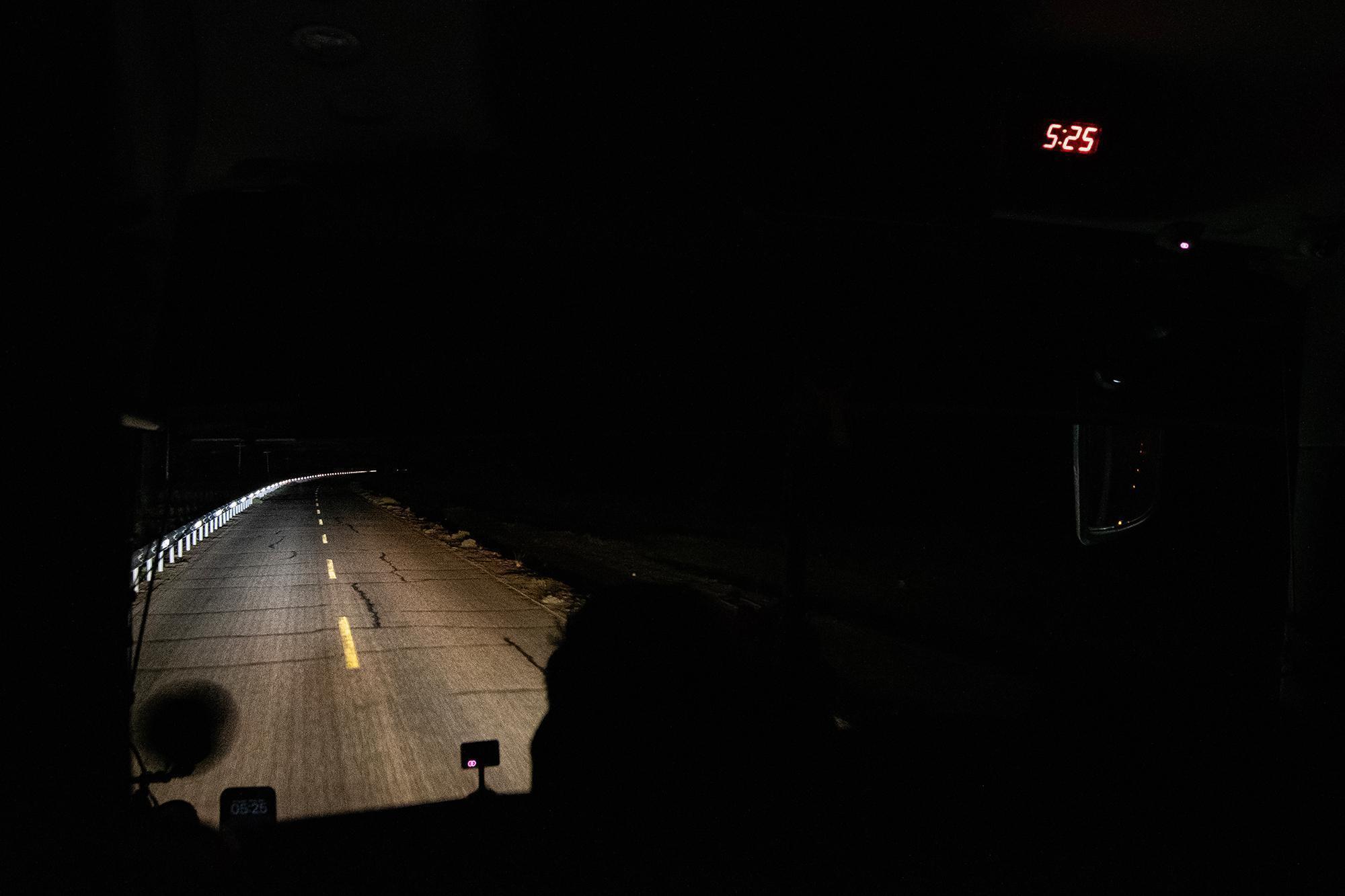 Interior view from the front of a bus at dawn, with only bus lights illuminating the road.