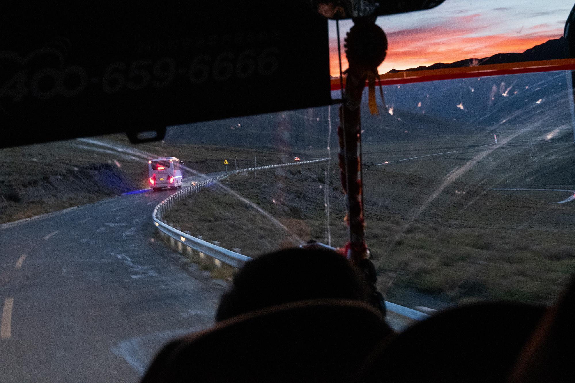 View from a bus at dusk, with another bus ahead on a winding road and an orange-red sunset behind the mountains.