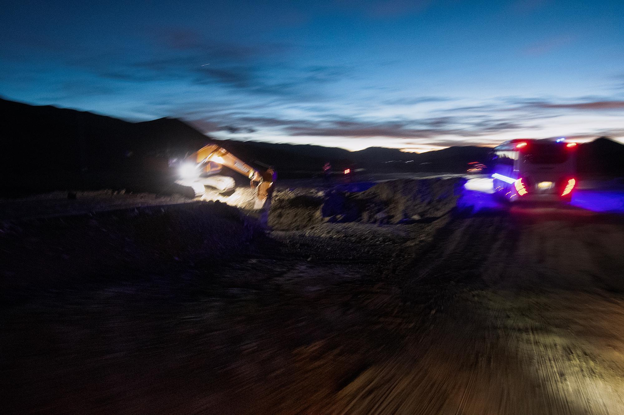Blurry construction scene at dusk, with an excavator and illuminated vehicle on a dirt road, with mountains in the background.