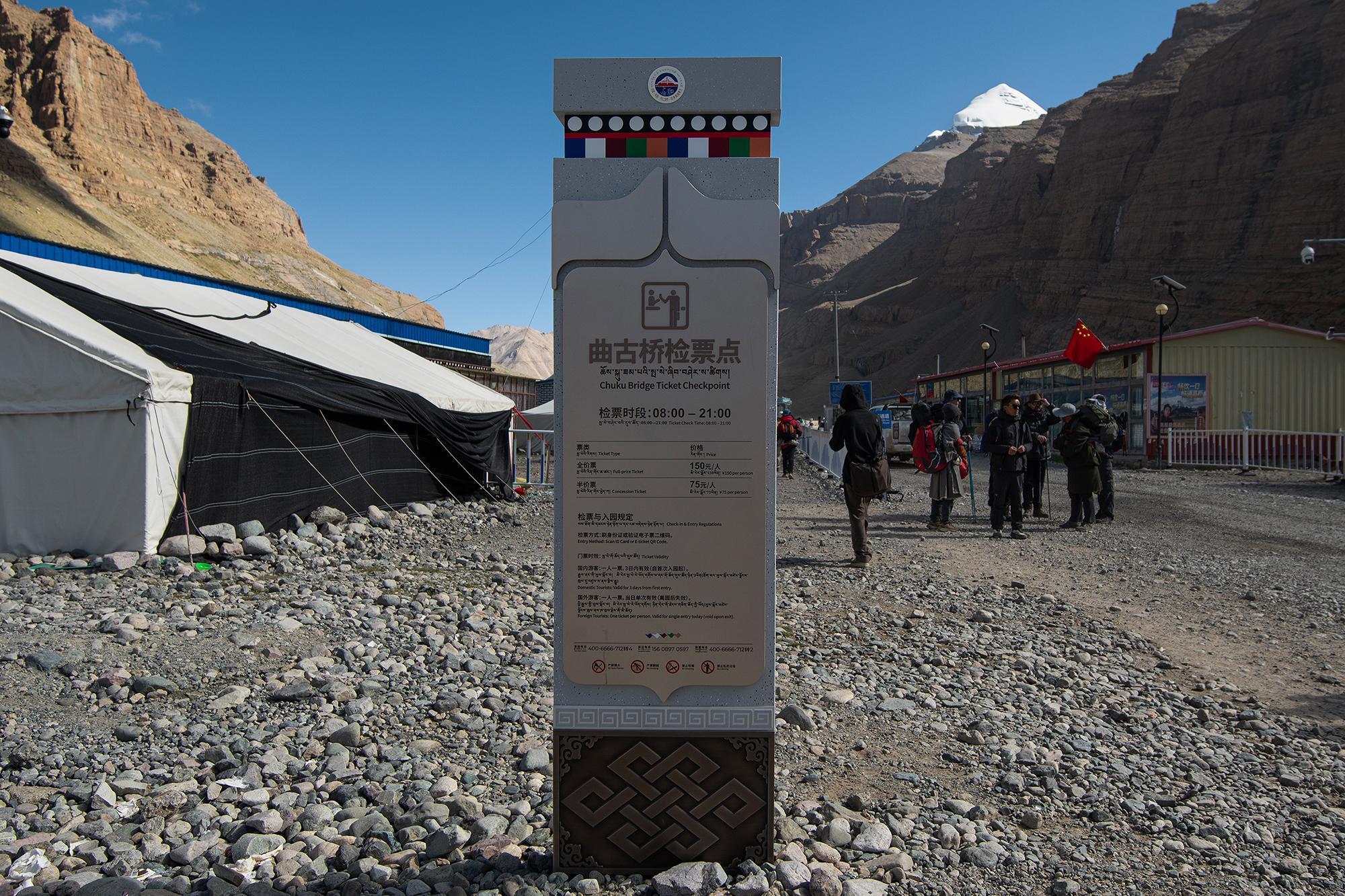 Chuku Bridge Ticket Checkpoint sign beside a rocky trail, with hikers, a building, and a snow-capped mountain in the background.
