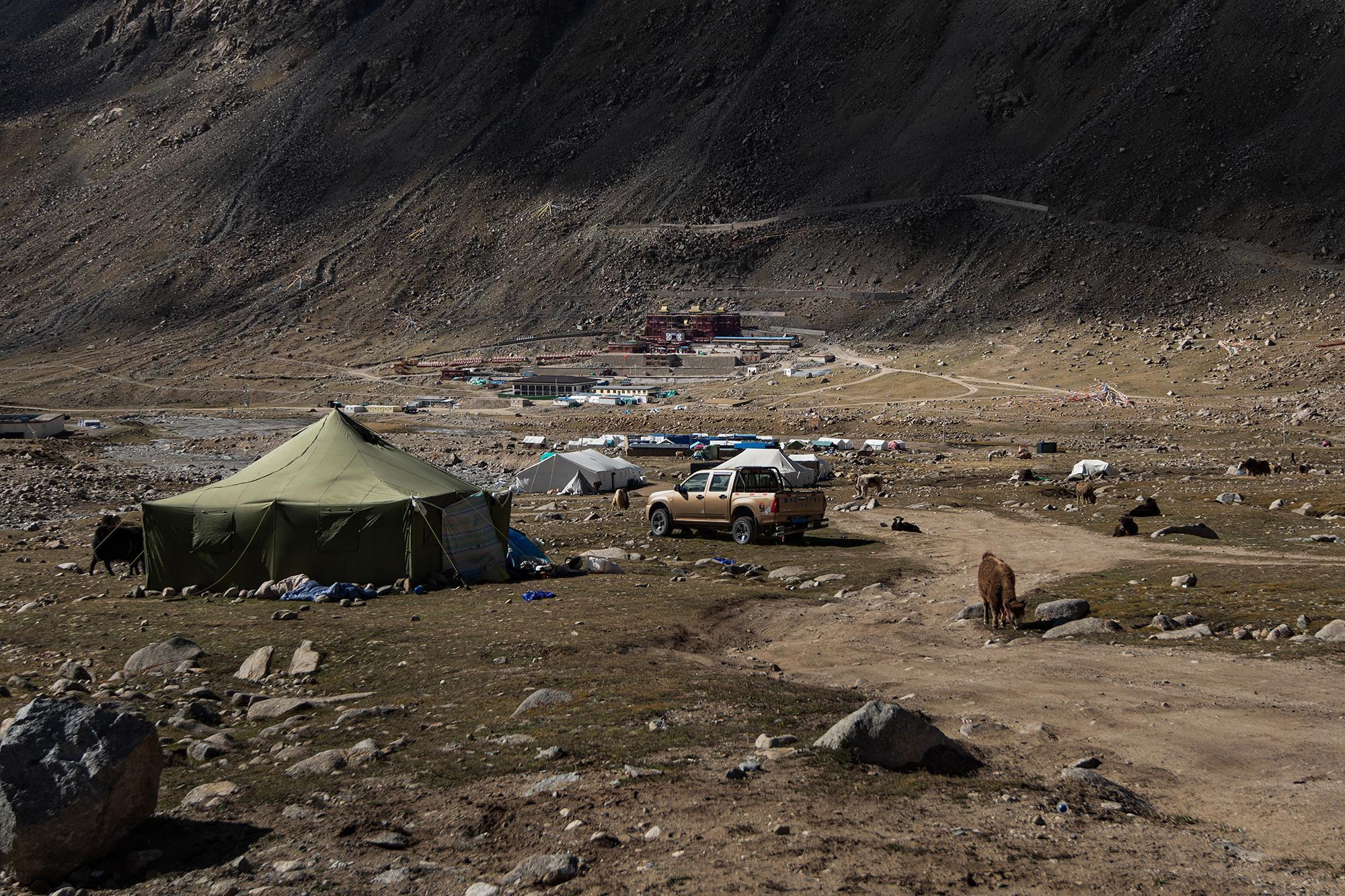 Temporary camp in a rocky valley, with tents, yaks, a pickup truck, and Dirapuk Monastery in the distance beneath towering mountains.