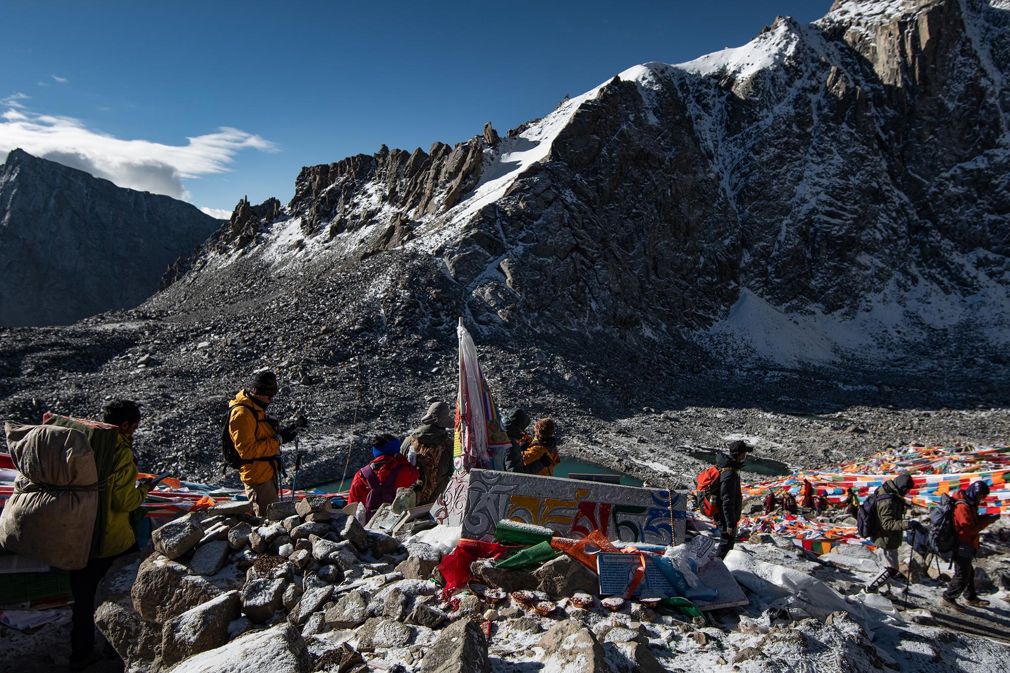 Hikers and porters crossing a snowy mountain pass, with a prayer-flag-covered cairn, turquoise glacial lake, and towering snow-capped peaks in the background.