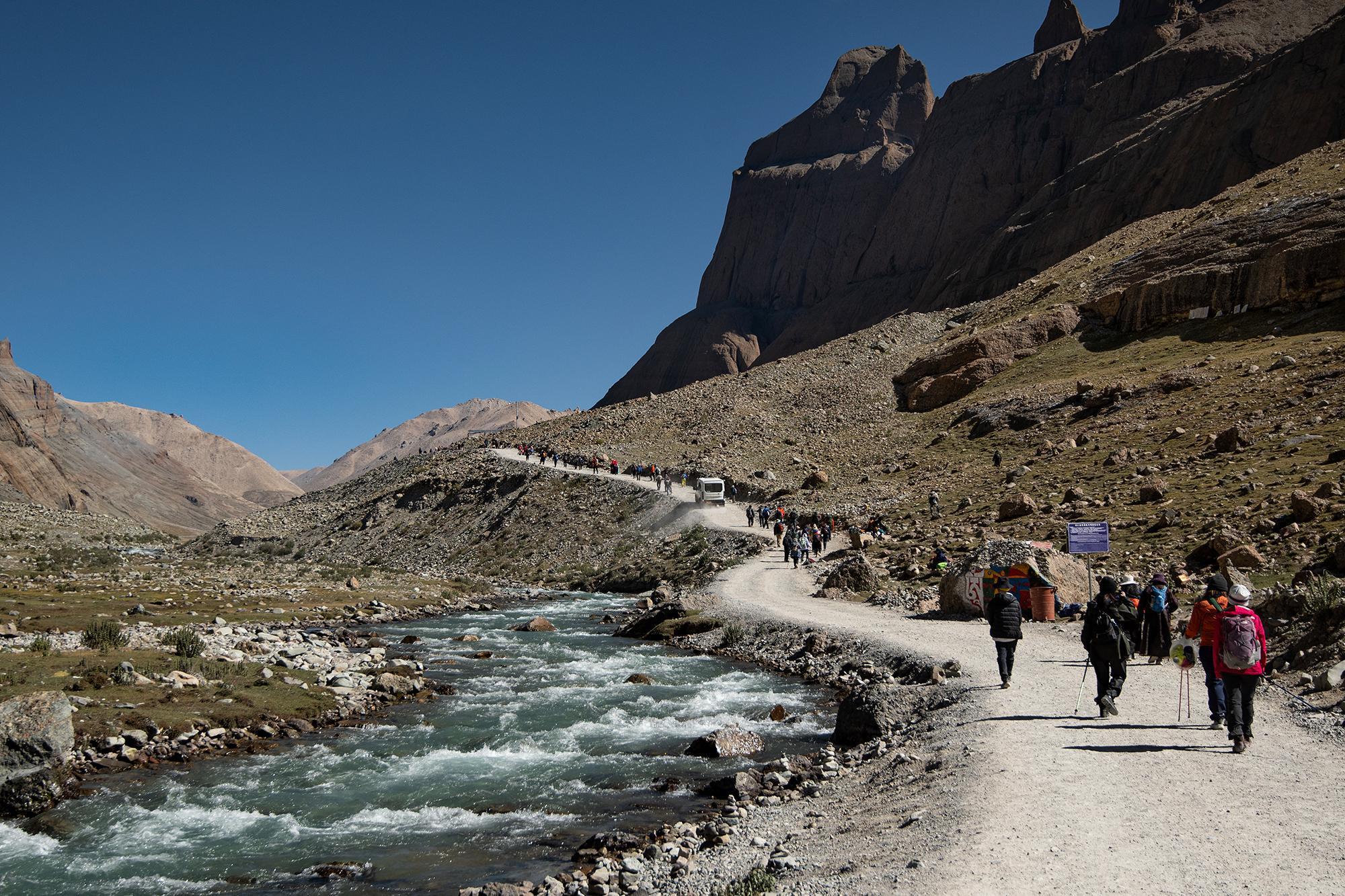 Hikers walking along a dirt road beside a fast-flowing river in a rugged mountain valley, with towering mountains under a clear blue sky.