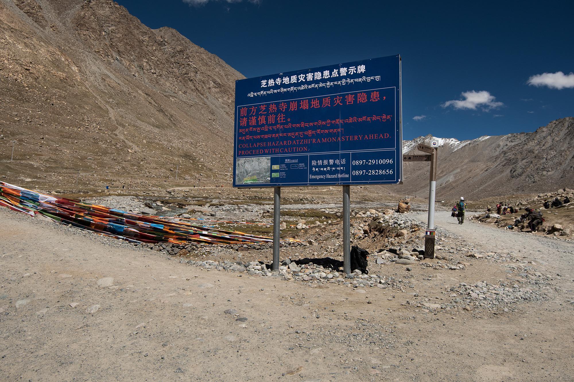 Blue warning sign for a collapse hazard at Zhi'ra Monastery, with prayer flags, a dirt path, and snow-capped mountains in the background.