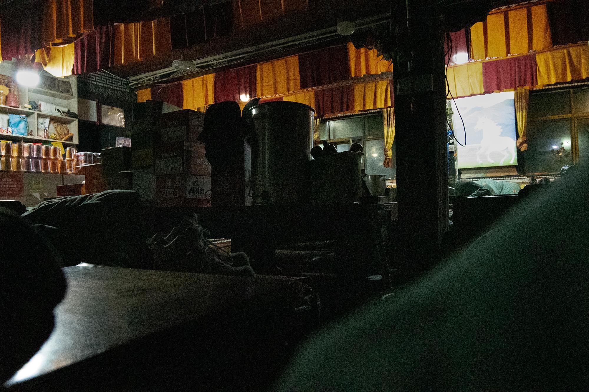 View from a tea house bench at night, with shelves of instant noodles, boxes, and metal containers in the dimly lit interior.