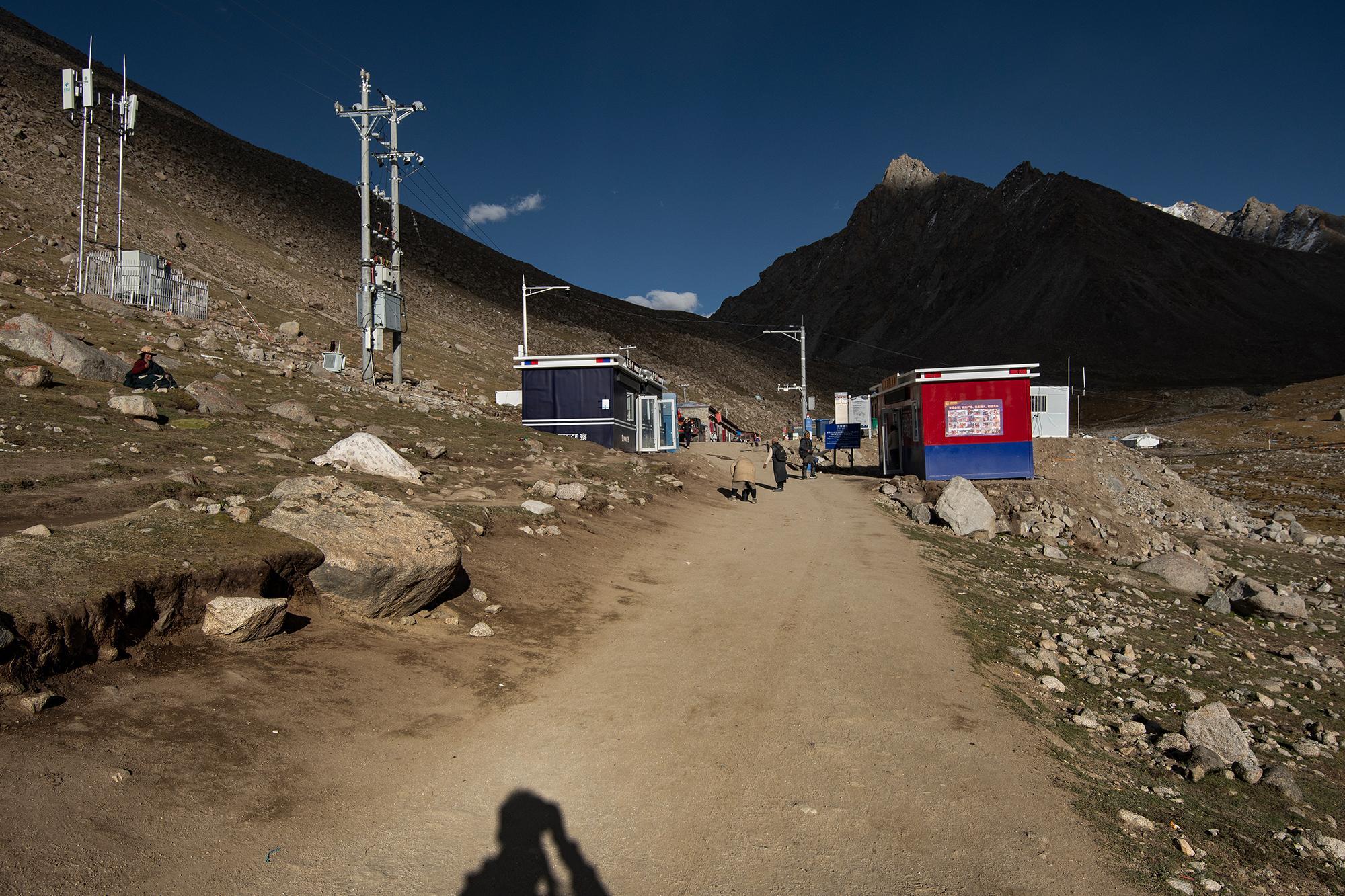 Rugged mountain landscape with a dirt path leading to a small settlement, surrounded by dark, snow-dusted mountains under a clear blue sky.