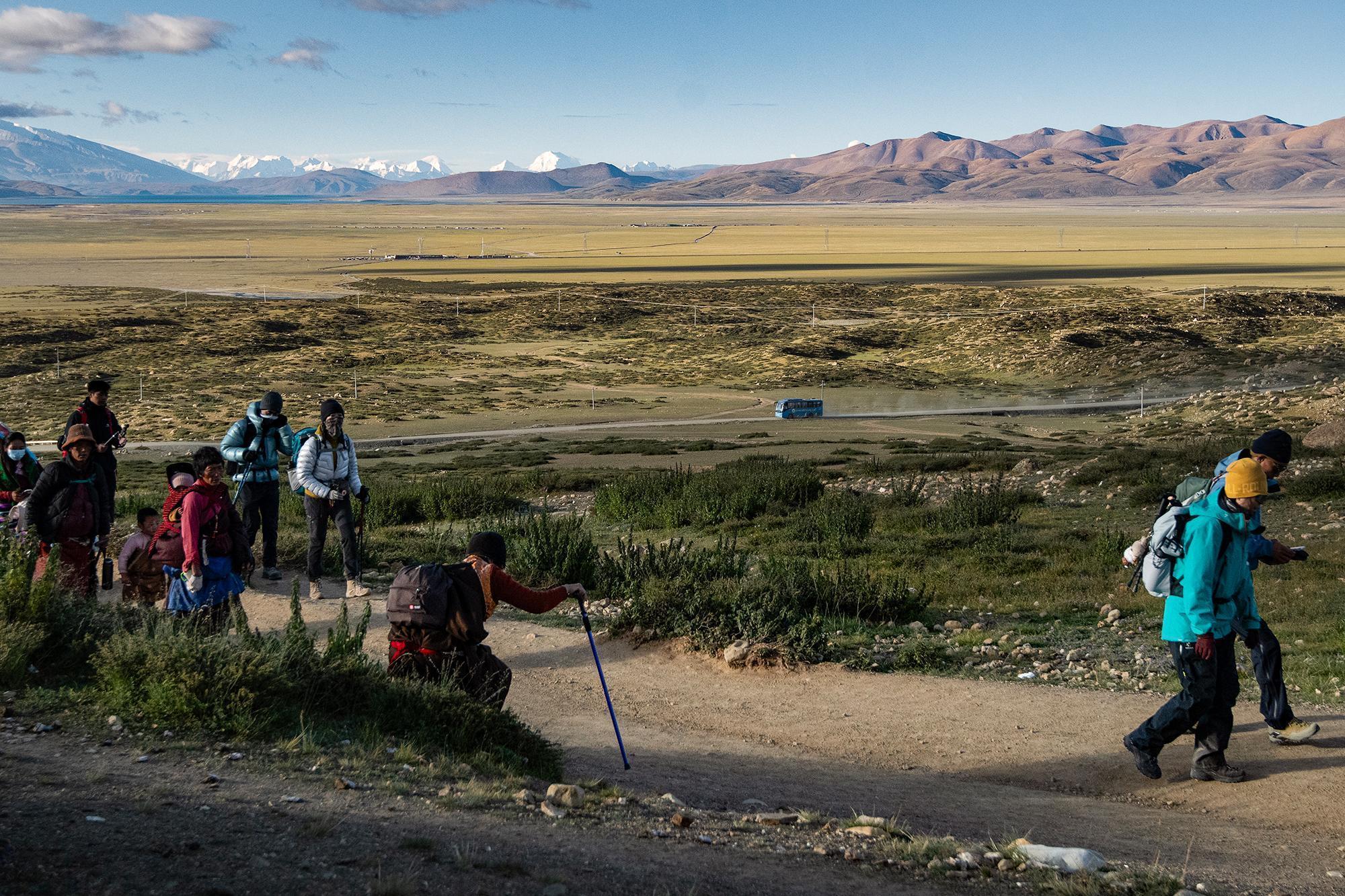 People trekking along a dirt path across a vast plain, with snow-capped mountains and a blue bus in the distance.