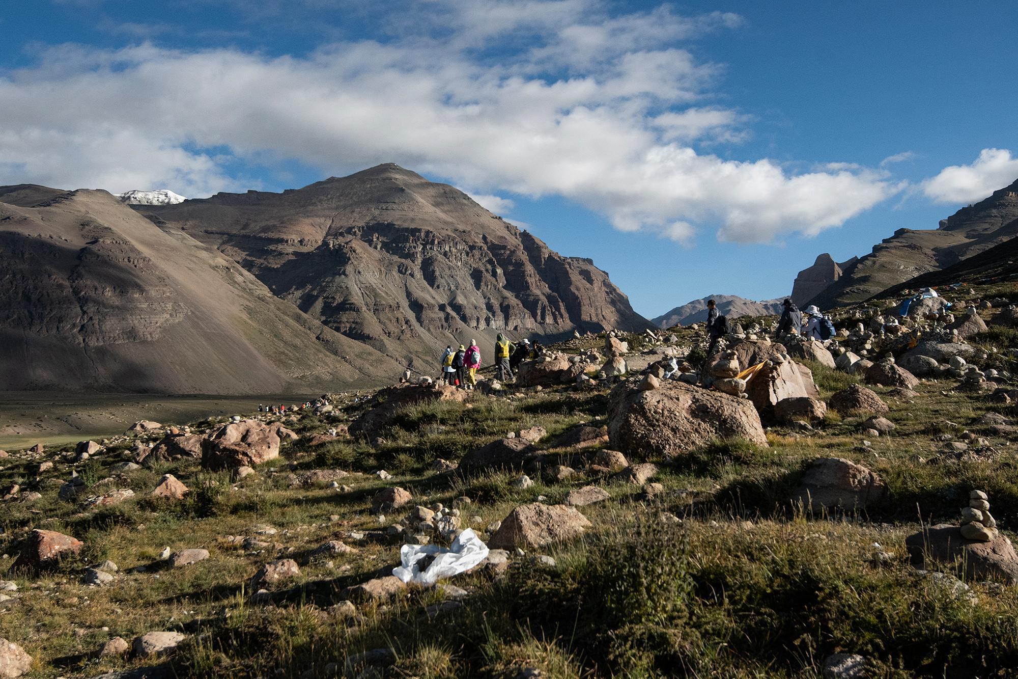 People trekking across a rocky hillside dotted with small rock cairns, with rugged mountains rising in the background.
