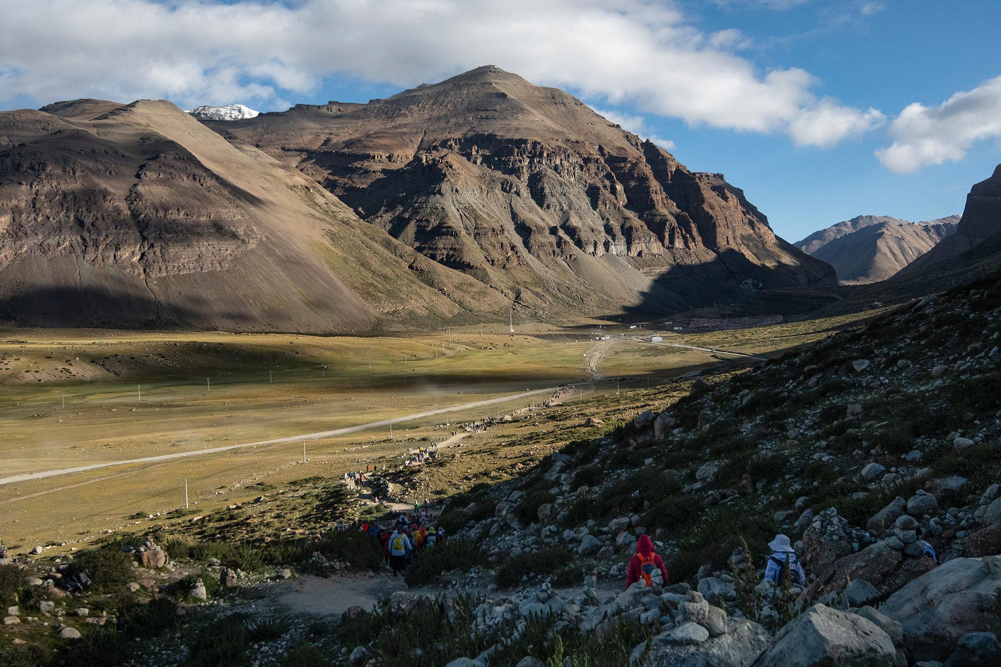 Hikers descending a rocky path above a grassy valley, with trekkers below and towering, rugged mountains in the background.