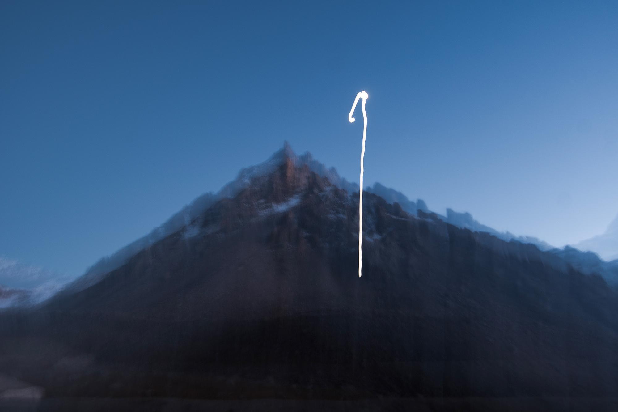 Long-exposure view of a dark mountain range beneath a deep blue sky, with a bright curved light trail across the scene.
