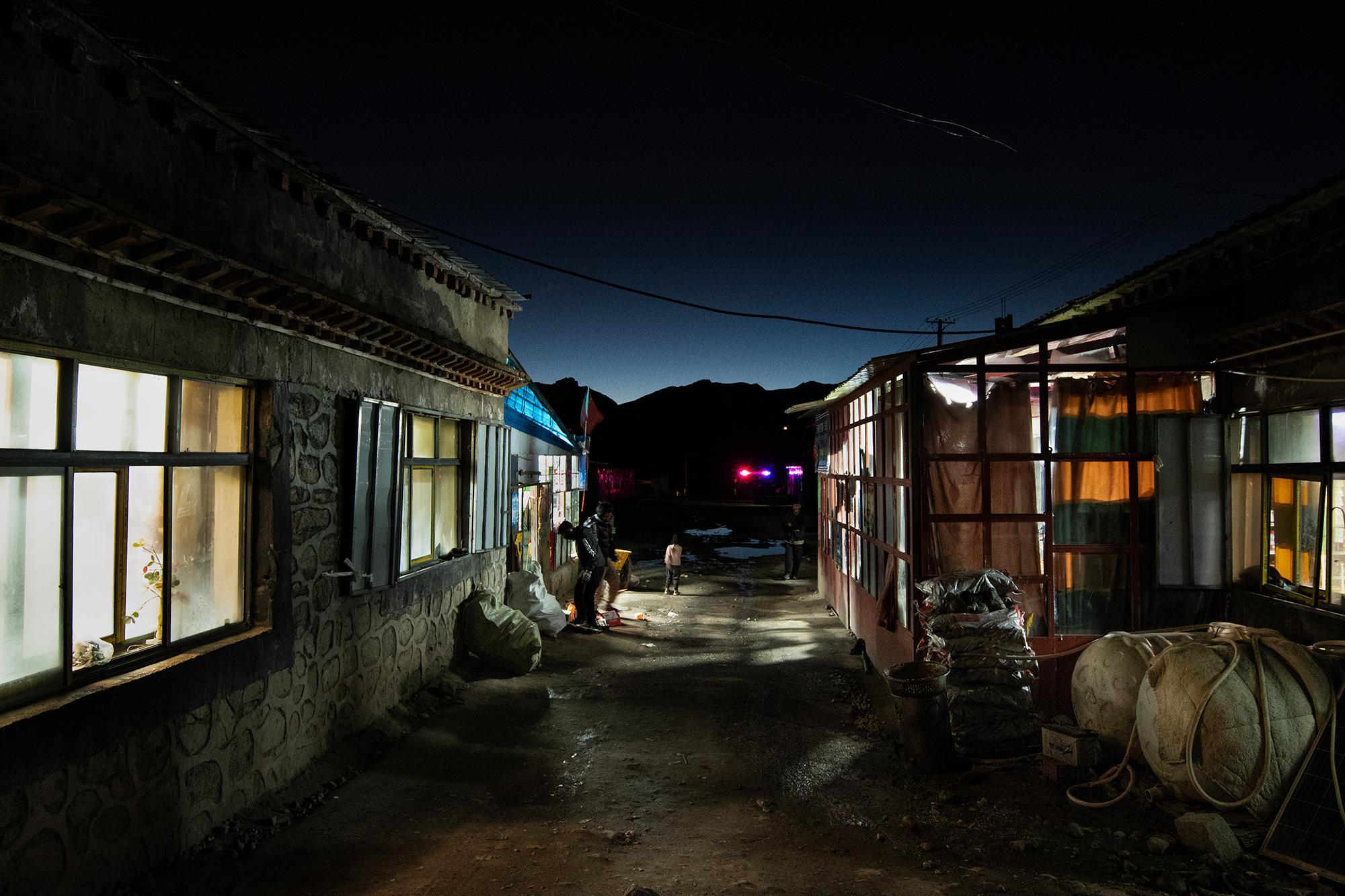 Dark alley between buildings at night, with lit windows, colorful tarps, a vehicle with flashing lights, and a few people in the distance.