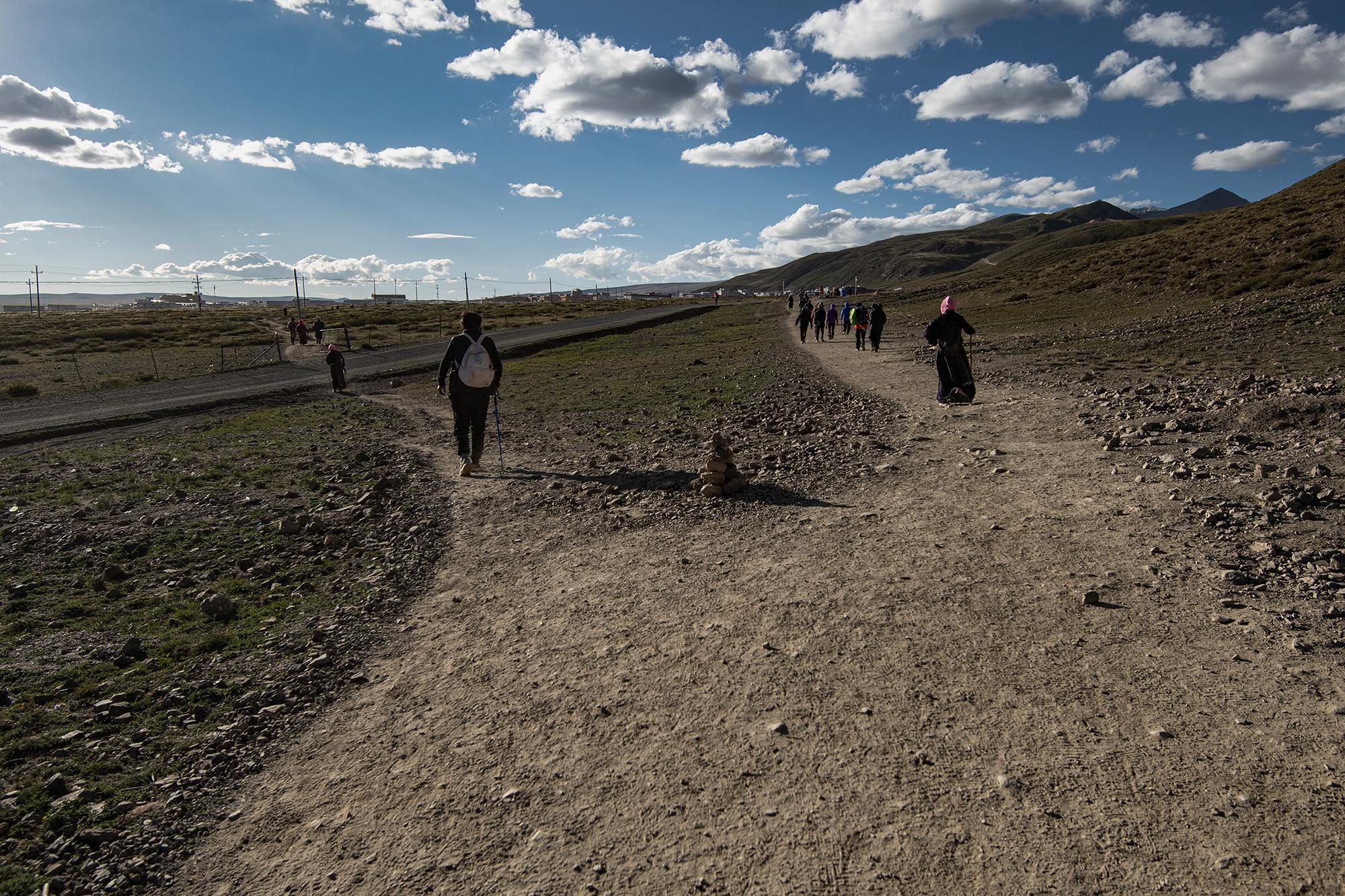 People walking along two diverging paths through an arid landscape, with a stack of stones between them and distant hills in the background.