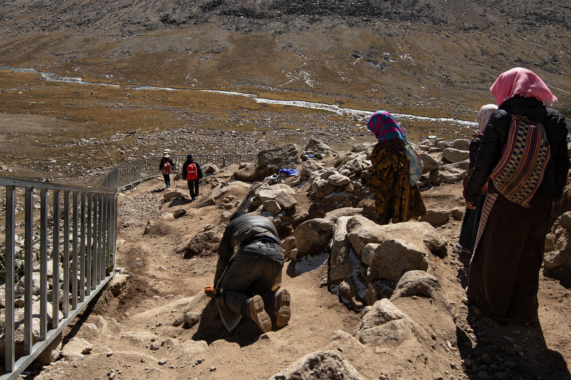 People on a rocky mountain path, with a kneeling pilgrim in the foreground, others walking along a metal railing, and a winding river below.