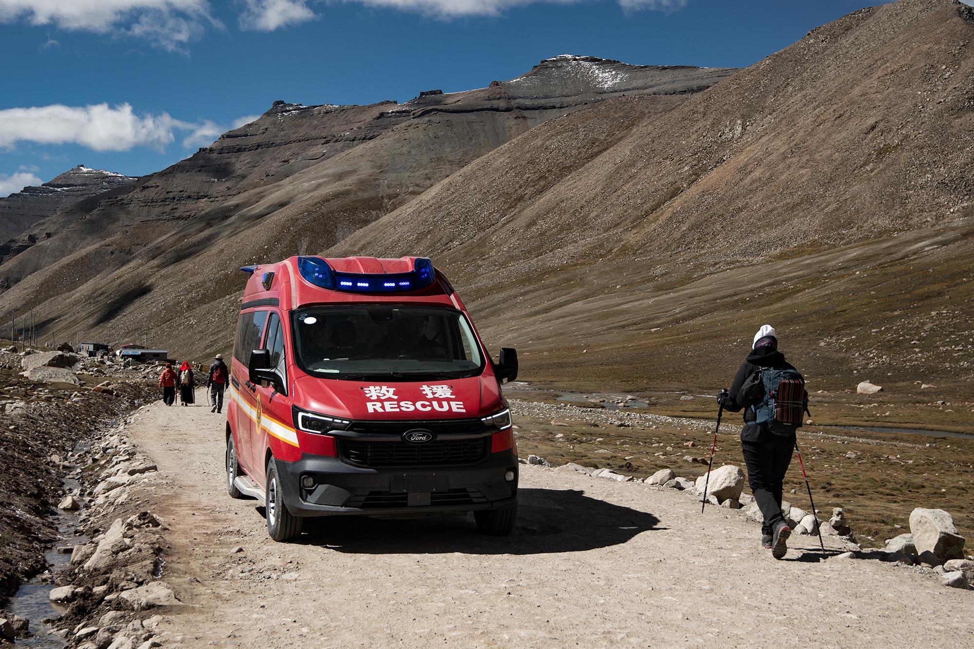 Red rescue van with flashing blue lights on a dirt road in an arid mountain landscape, with hikers and small buildings in the background.