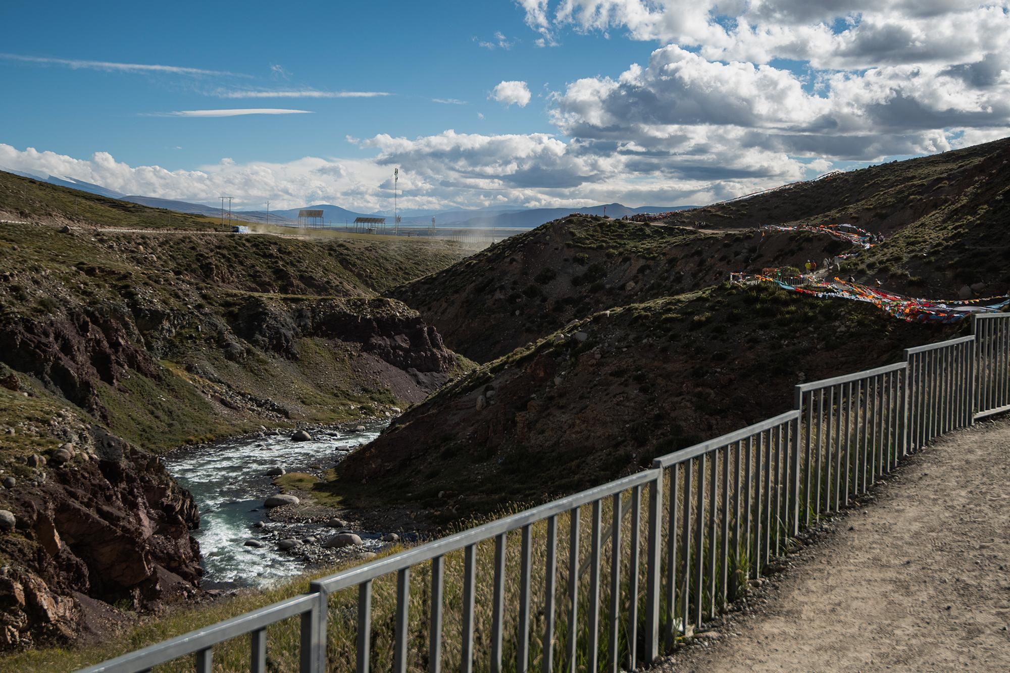 Winding river through a rocky valley, with a dirt path, prayer flags and a distant road in the background.