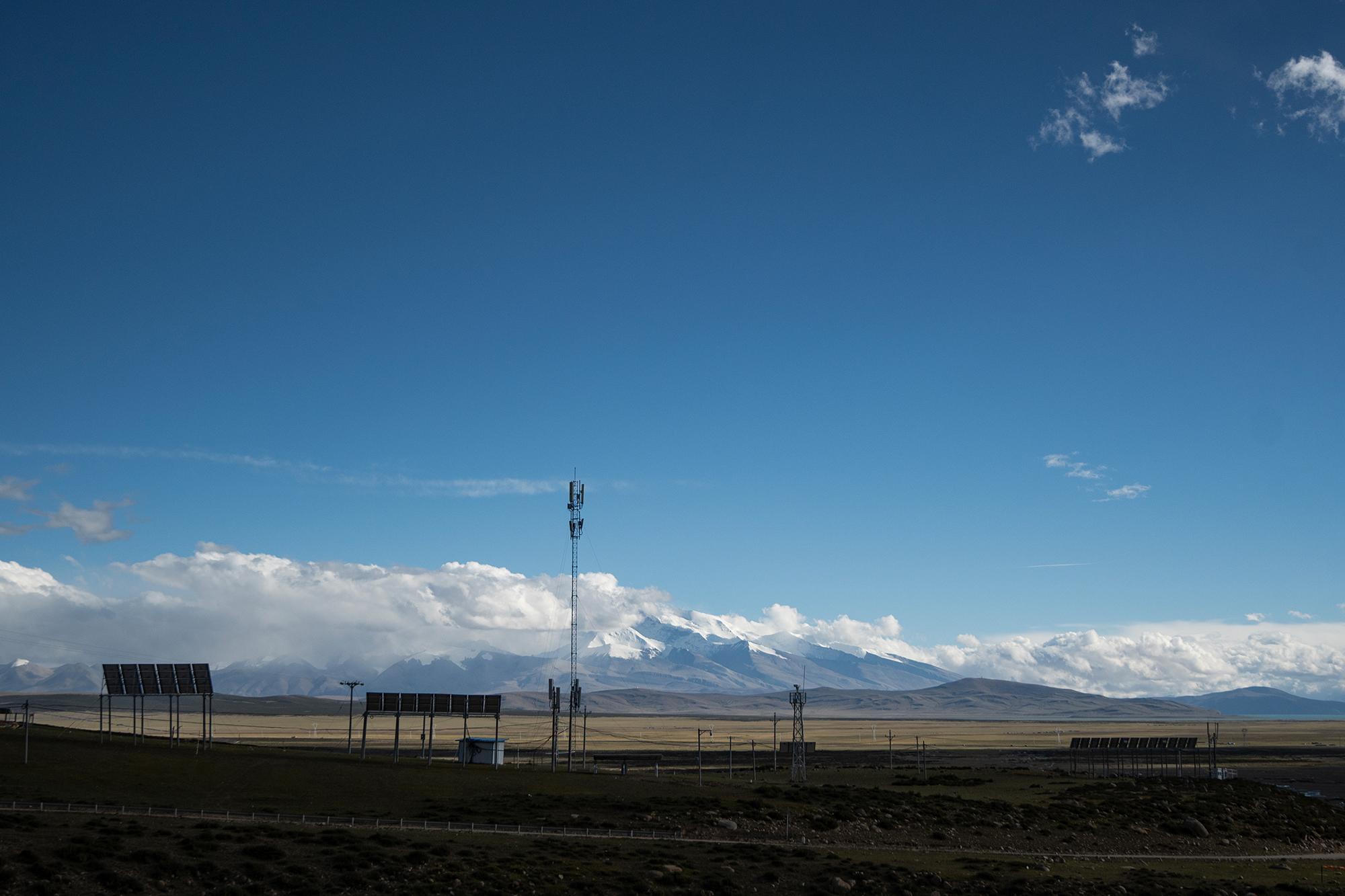 Open scrubland with solar panels and cellular towers, stretching toward a golden plain and snow-capped mountains under a blue sky.