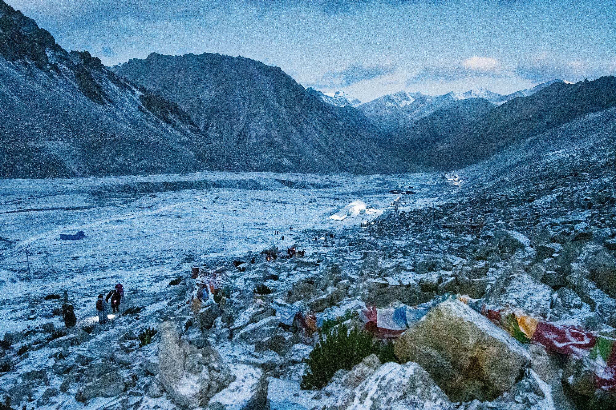 Trekkers with headlamps walking through a snowy mountain valley at dawn, with prayer flags, lit tents, and snow-dusted peaks in the background.