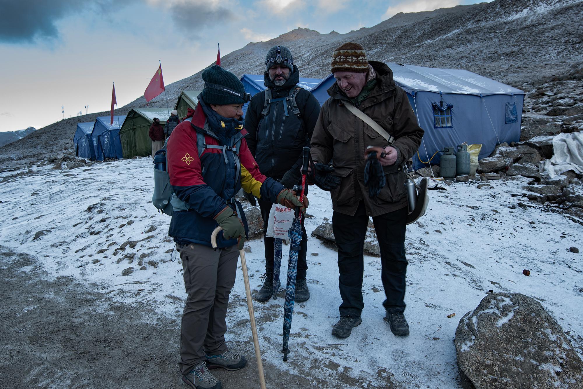 Three hikers at a snow-dusted high-altitude camp, gathered around a plaid umbrella, with tents and Chinese flags in the background.