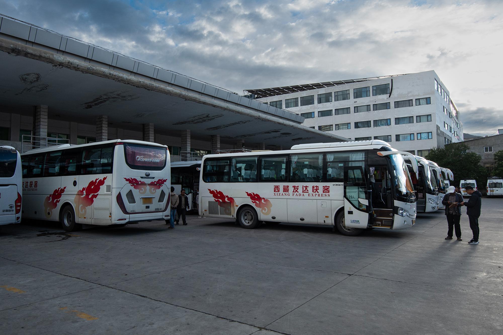 White buses parked at a bus station, with two men standing nearby and a large building in the background.