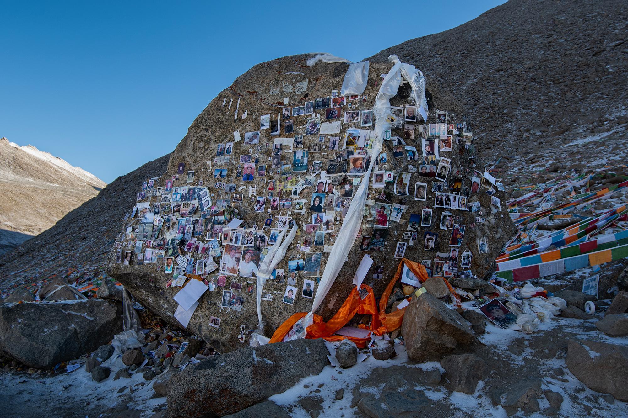 A boulder covered in photographs, draped with white and orange prayer flags on a snowy mountain slope.