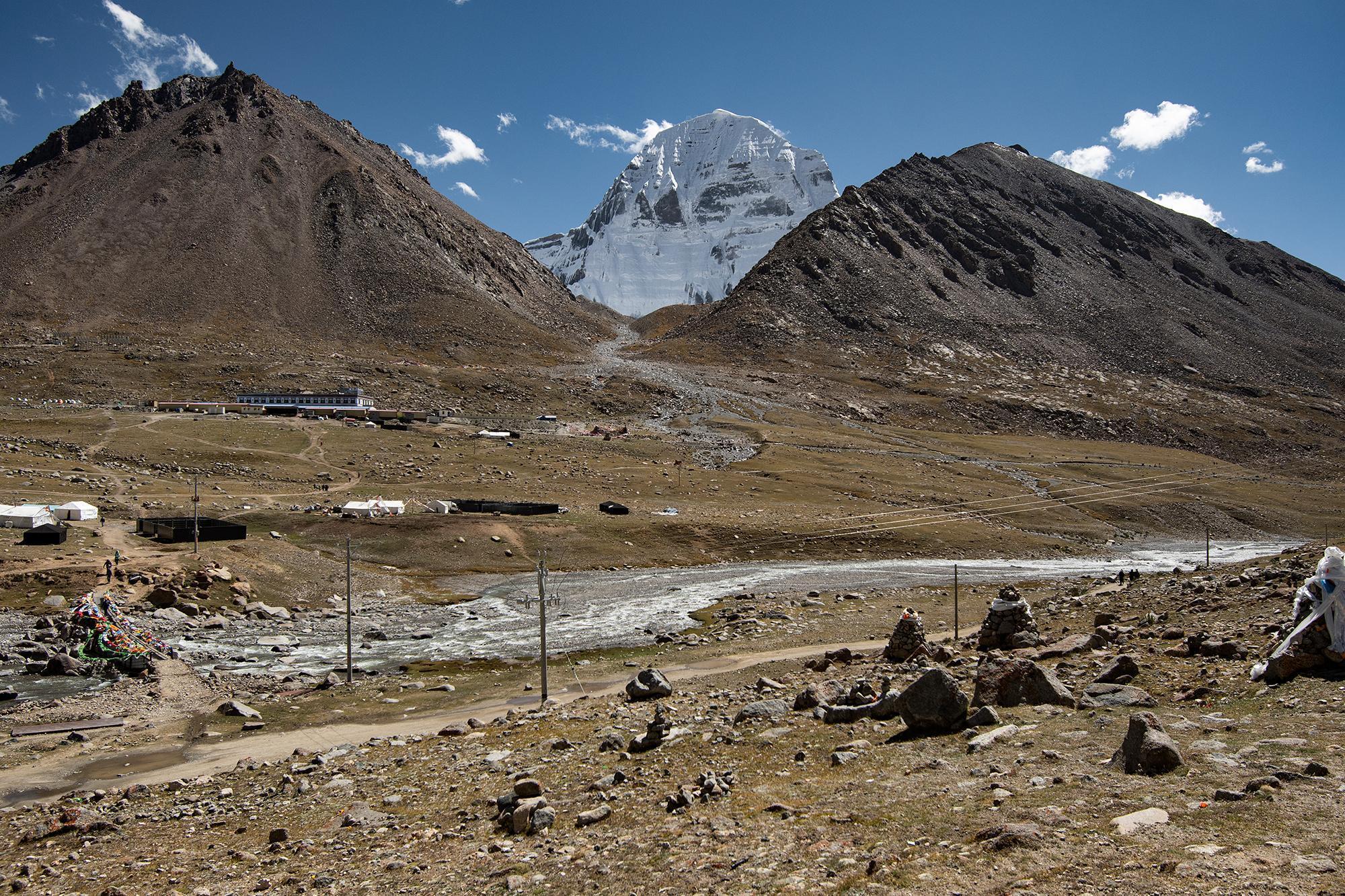 Mount Kailash rising above a wide valley, with a winding river, dirt road, buildings, prayer flags, and rocky terrain in the foreground.