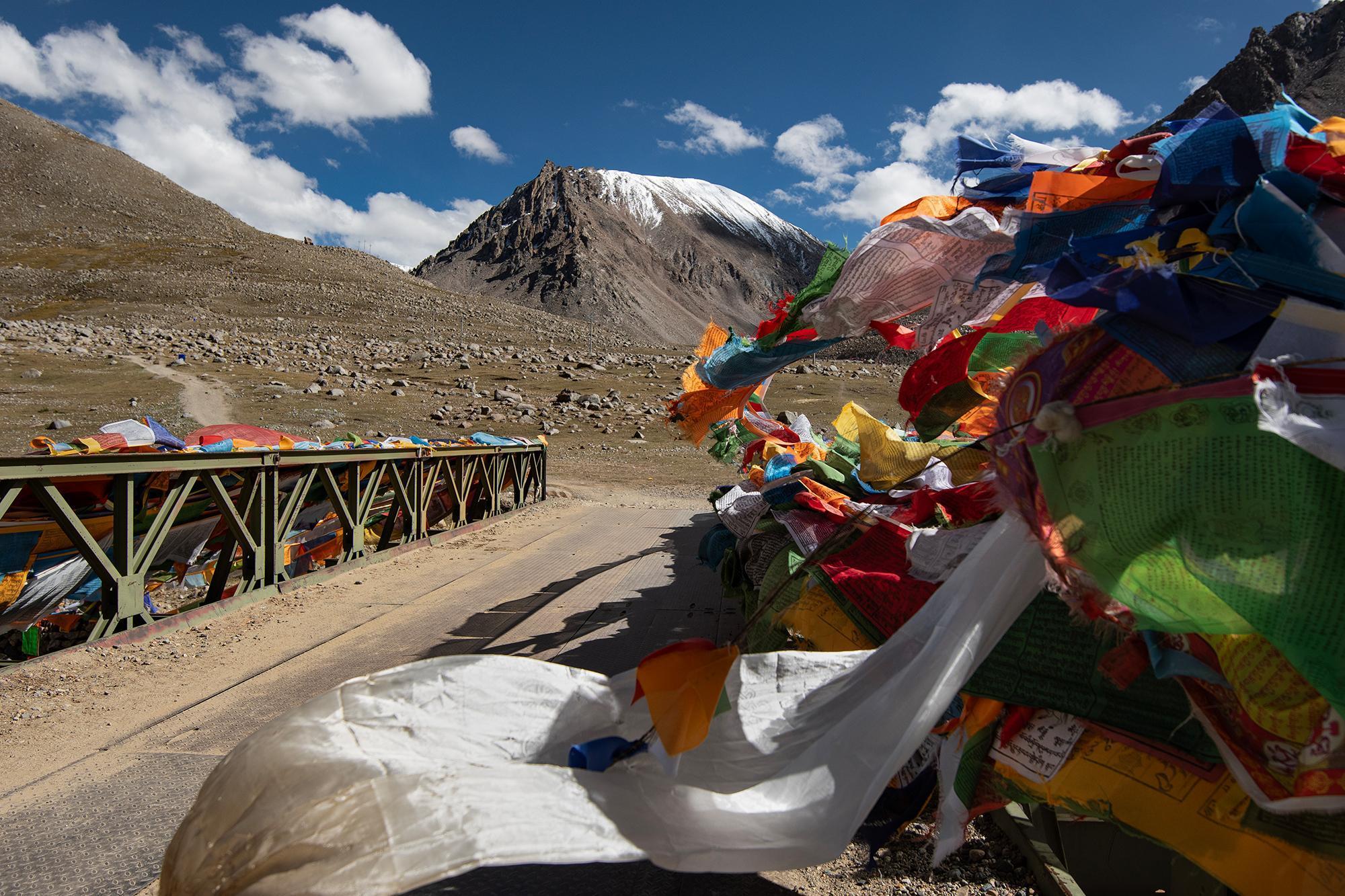 Colorful prayer flags along a bridge, with a dirt road leading toward a rocky mountain landscape and snow-capped peak.