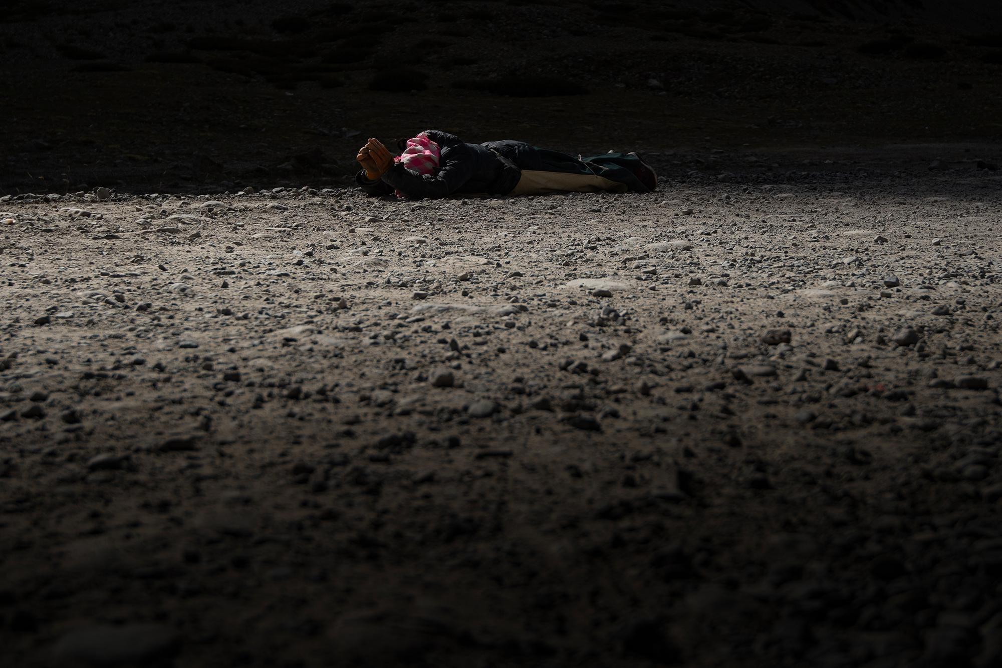 A pilgrim prostrating on rocky ground, with sunlight falling across the scene and deep shadows surrounding them.