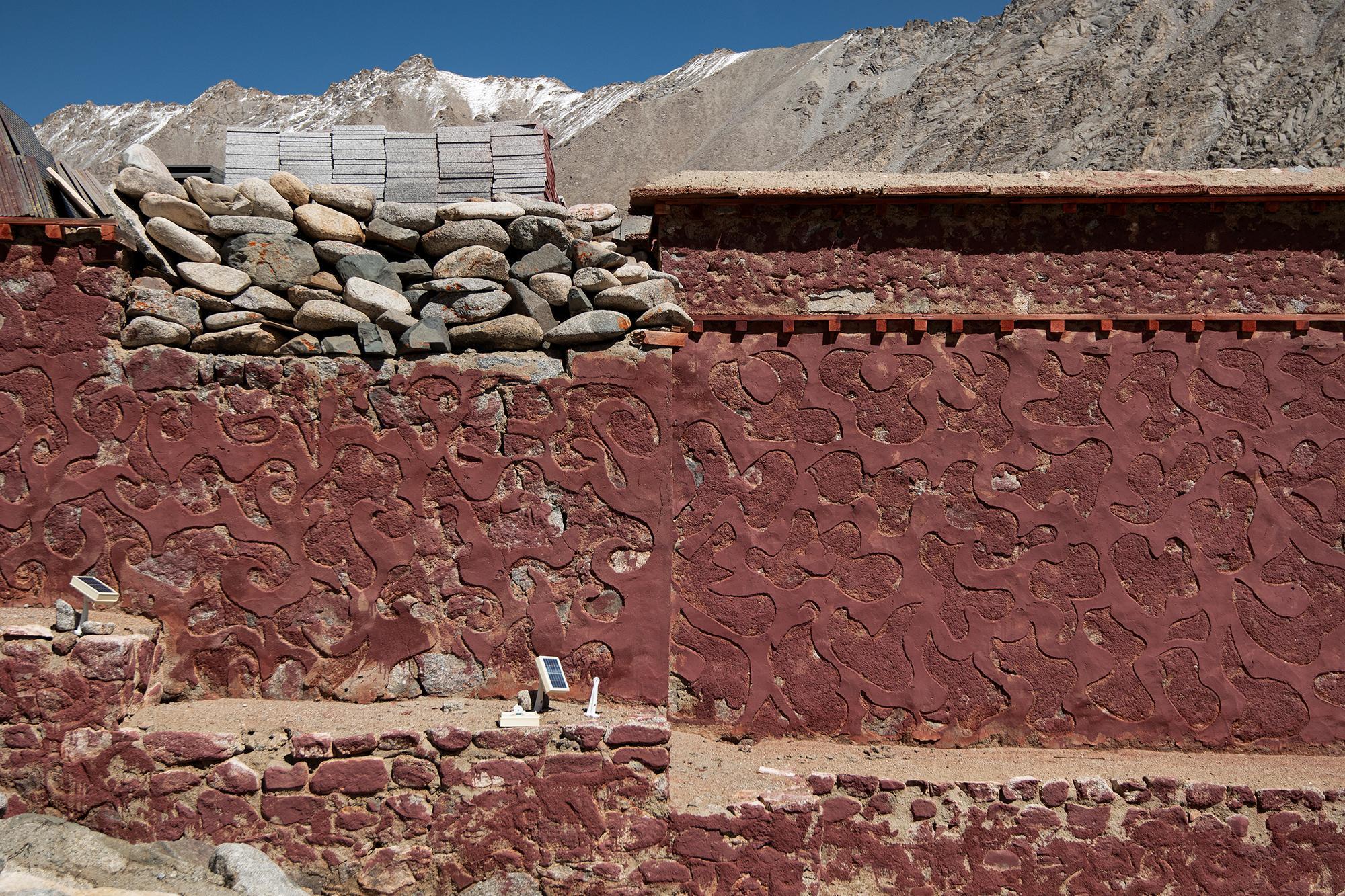 A vibrant red wall with carved patterns, topped by rough stone and grey bricks, with snow-capped mountains in the background.