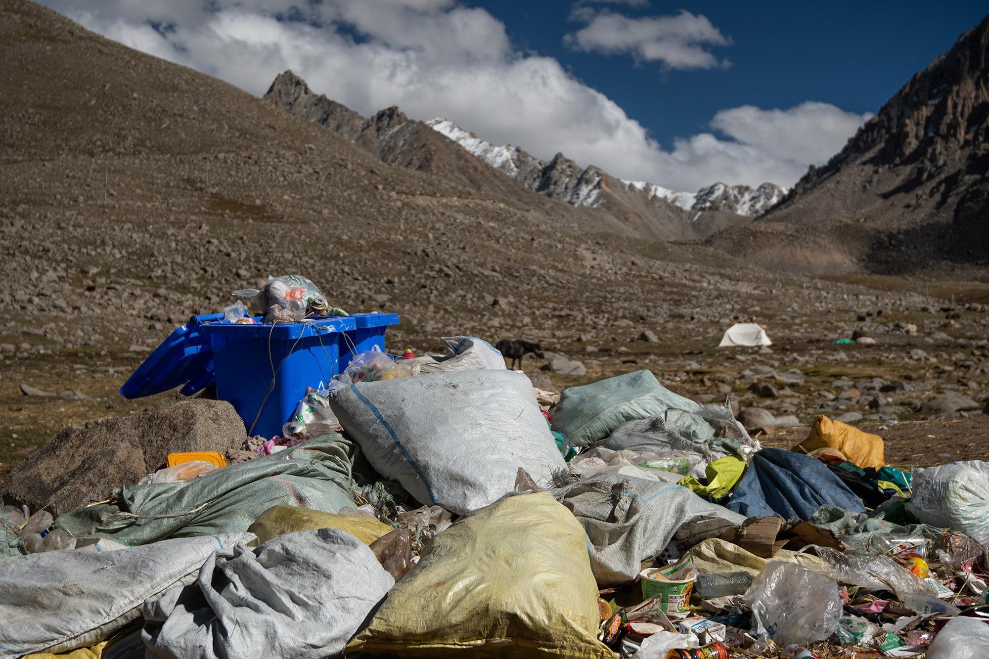 A rocky mountain valley with an overflowing dumpster and piles of trash in the foreground, with snow-capped peaks in the background.
